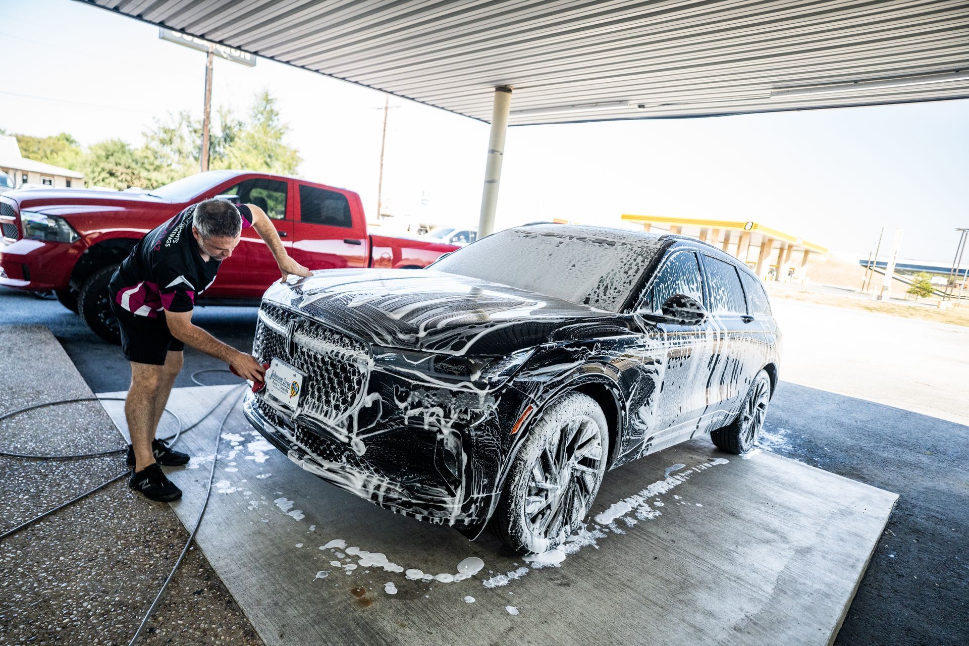 Man washing a black SUV covered in soap suds at a car wash; a red truck is parked nearby.