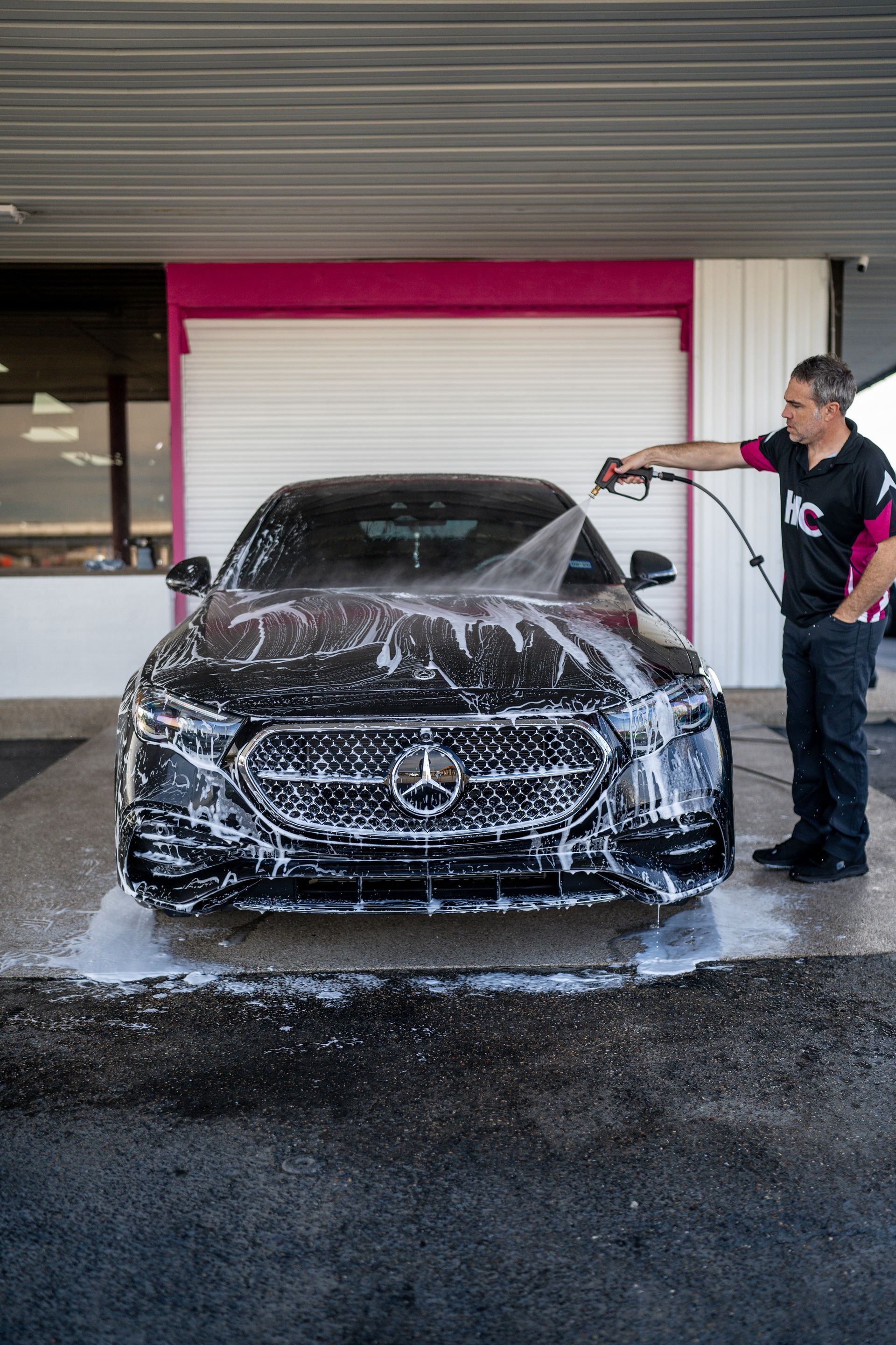 Man washing a black Mercedes with a spray hose at a car wash; soapy water covers the car.