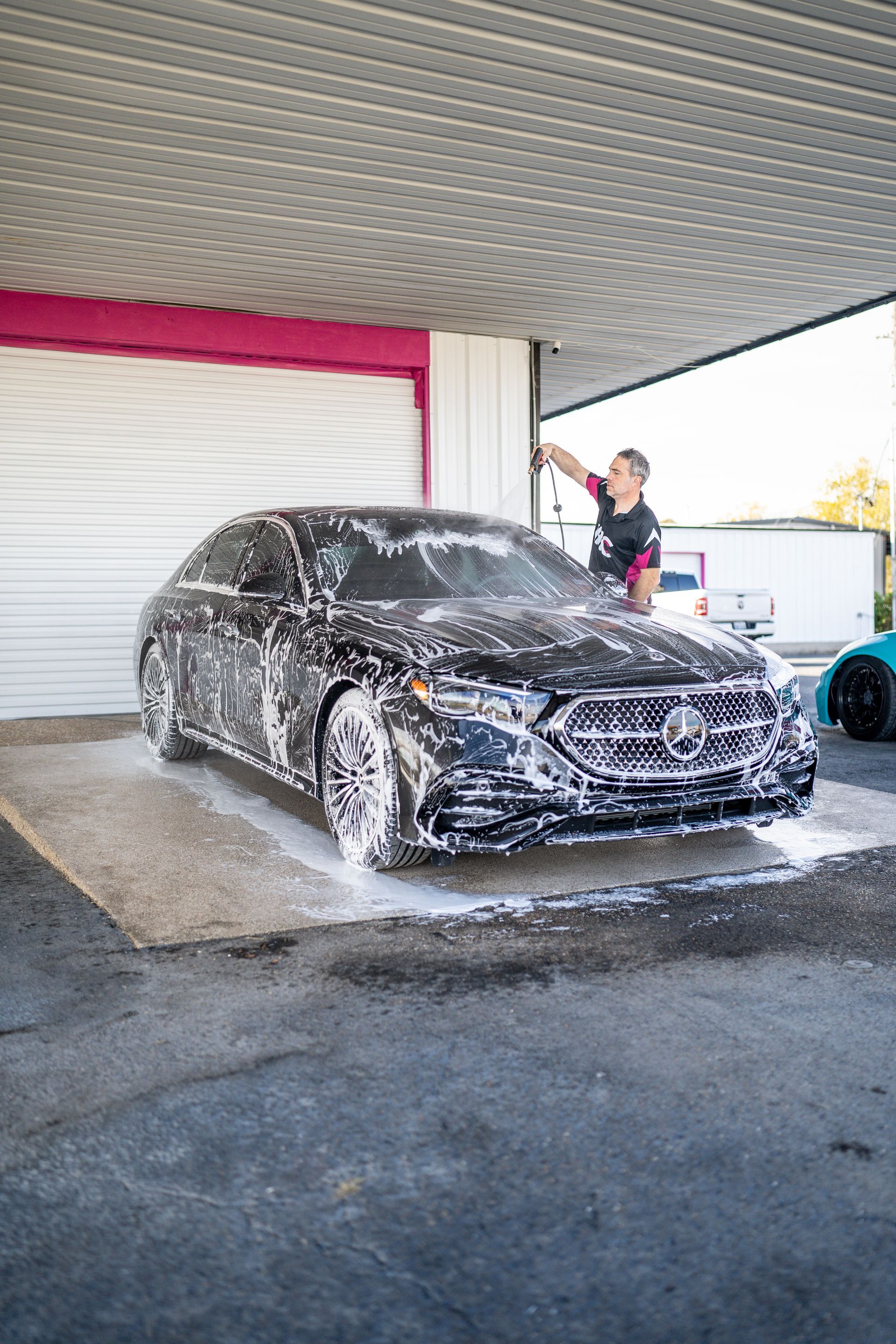 Person washing a black Mercedes sedan at a car wash. White foam covers the car.