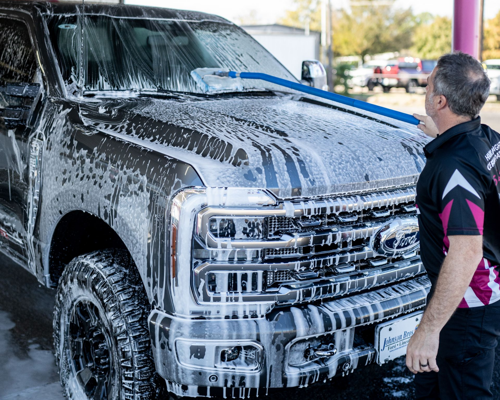 Man washing a dark pickup truck with a blue brush, covered in soap at a car wash.