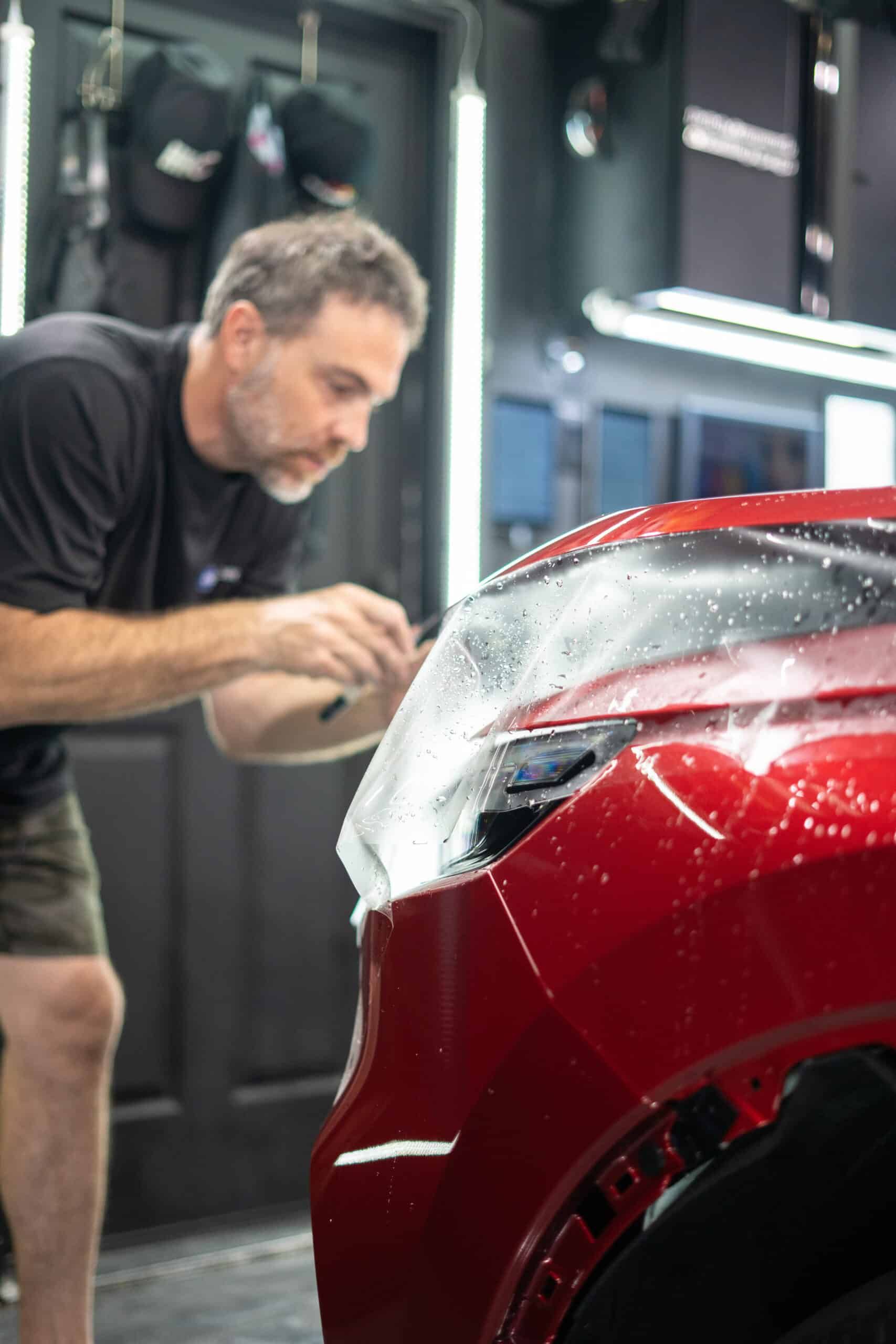 A man is applying a protective film to the front of a red car.