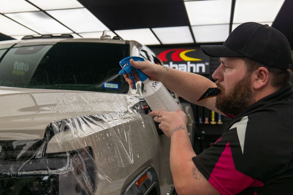 Man spraying a solution on a car's hood, preparing it for a protective film application in a shop.
