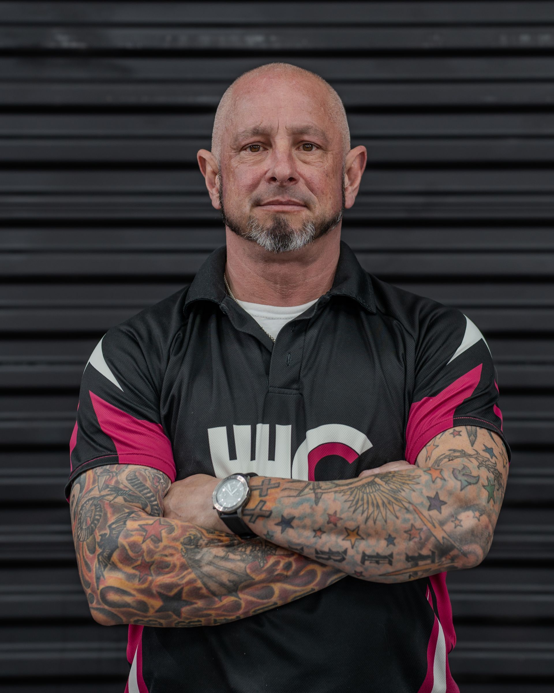 Man with tattoos and crossed arms, wearing a black shirt with pink accents in front of a dark background.