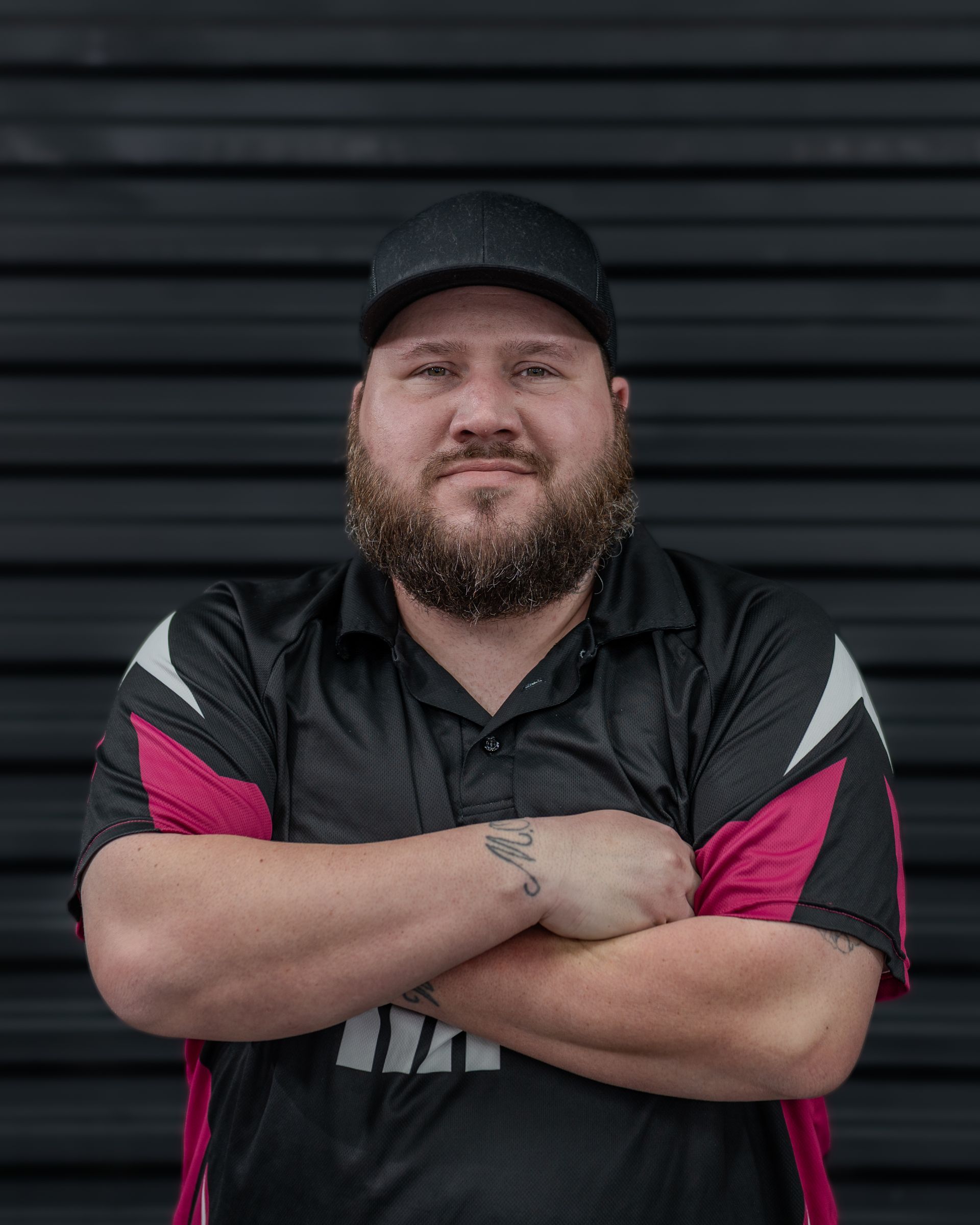 Man with beard and hat, arms crossed, wearing a black and pink shirt, in front of a dark background.