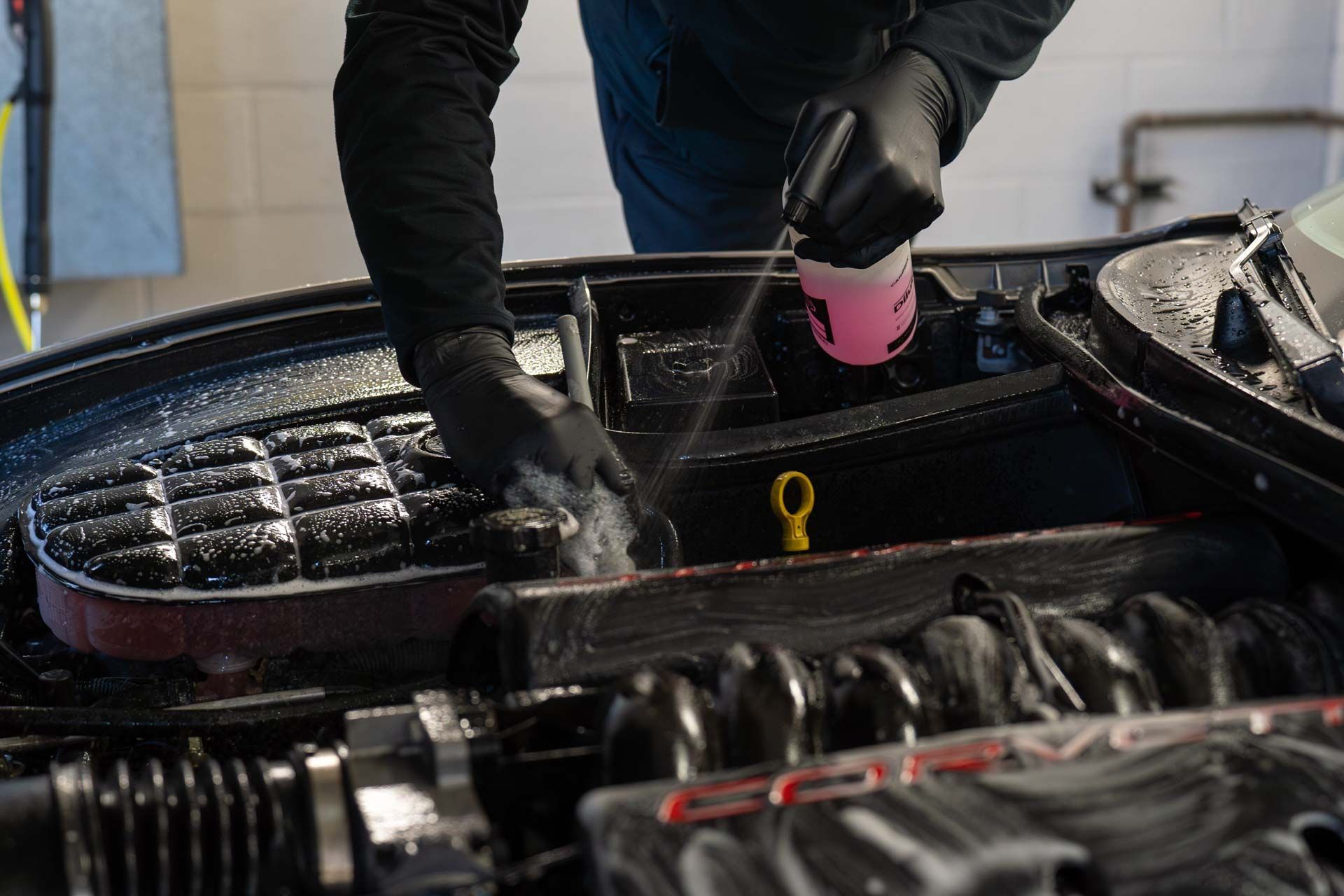 Person cleaning a car engine with a spray bottle, wearing gloves in a shop.