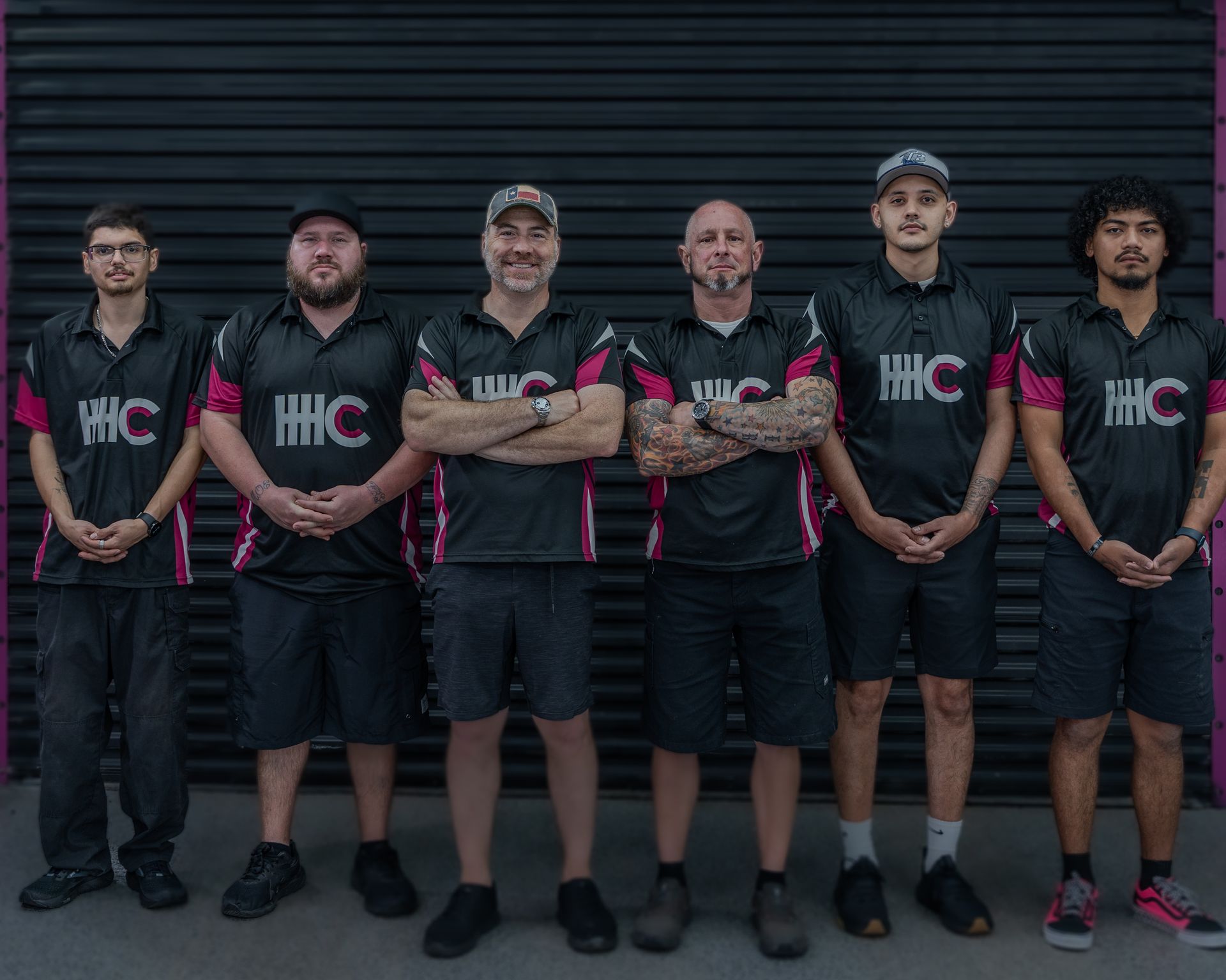 Group of seven people wearing matching black shirts with pink accents, standing in front of a dark wall.