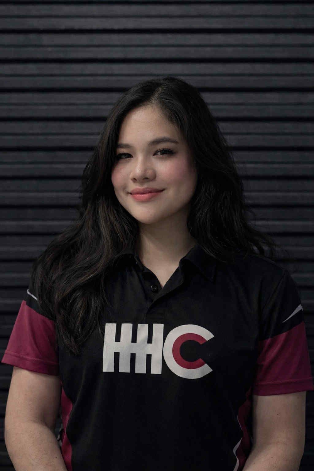 Woman in black and pink shirt smiling; IHC logo on shirt, against a dark, ridged wall.