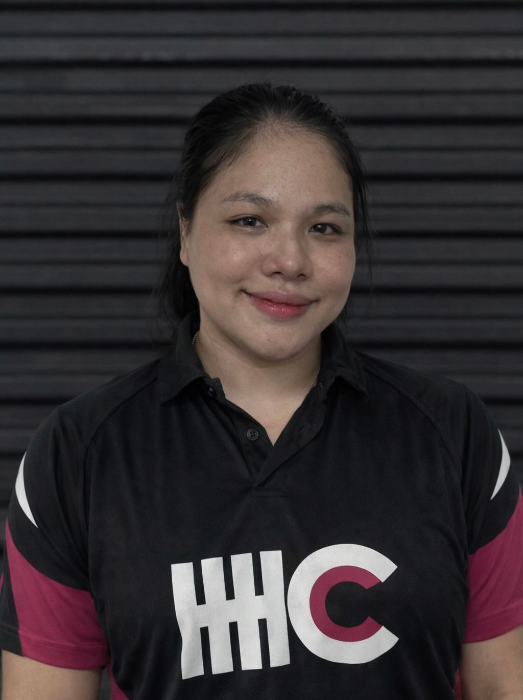 Woman in black and pink shirt smiling; IHC logo on shirt, against a dark, ridged wall.