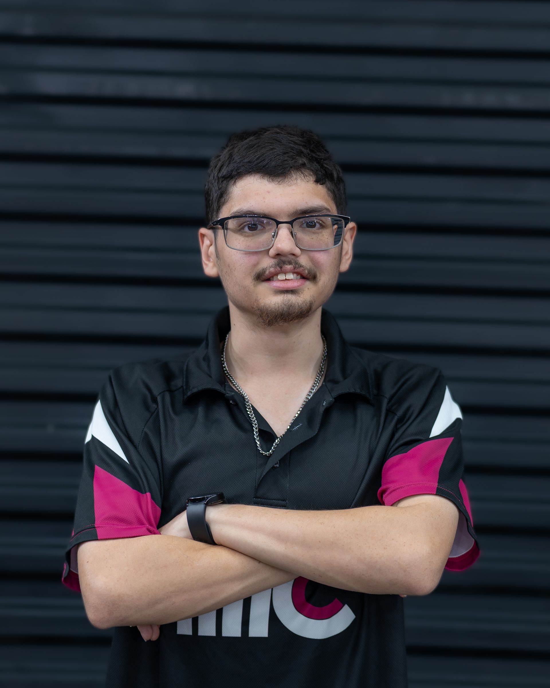 Man wearing glasses and a black and pink shirt, arms crossed, smiling. Dark background.