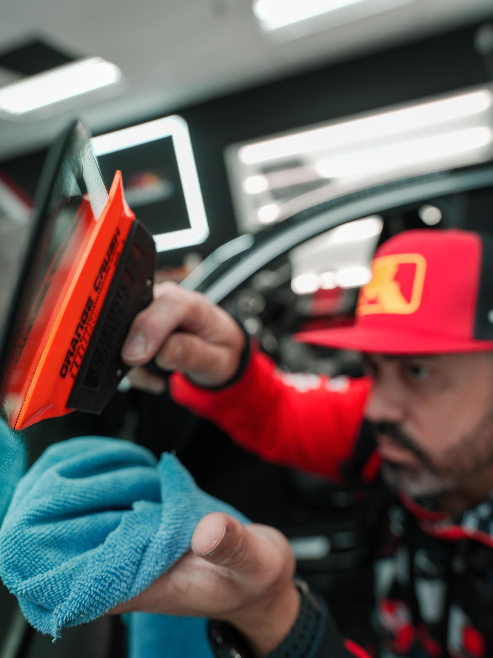 Man installing car window tint, using a squeegee and microfiber cloth. Wearing red cap and jacket, in a shop.