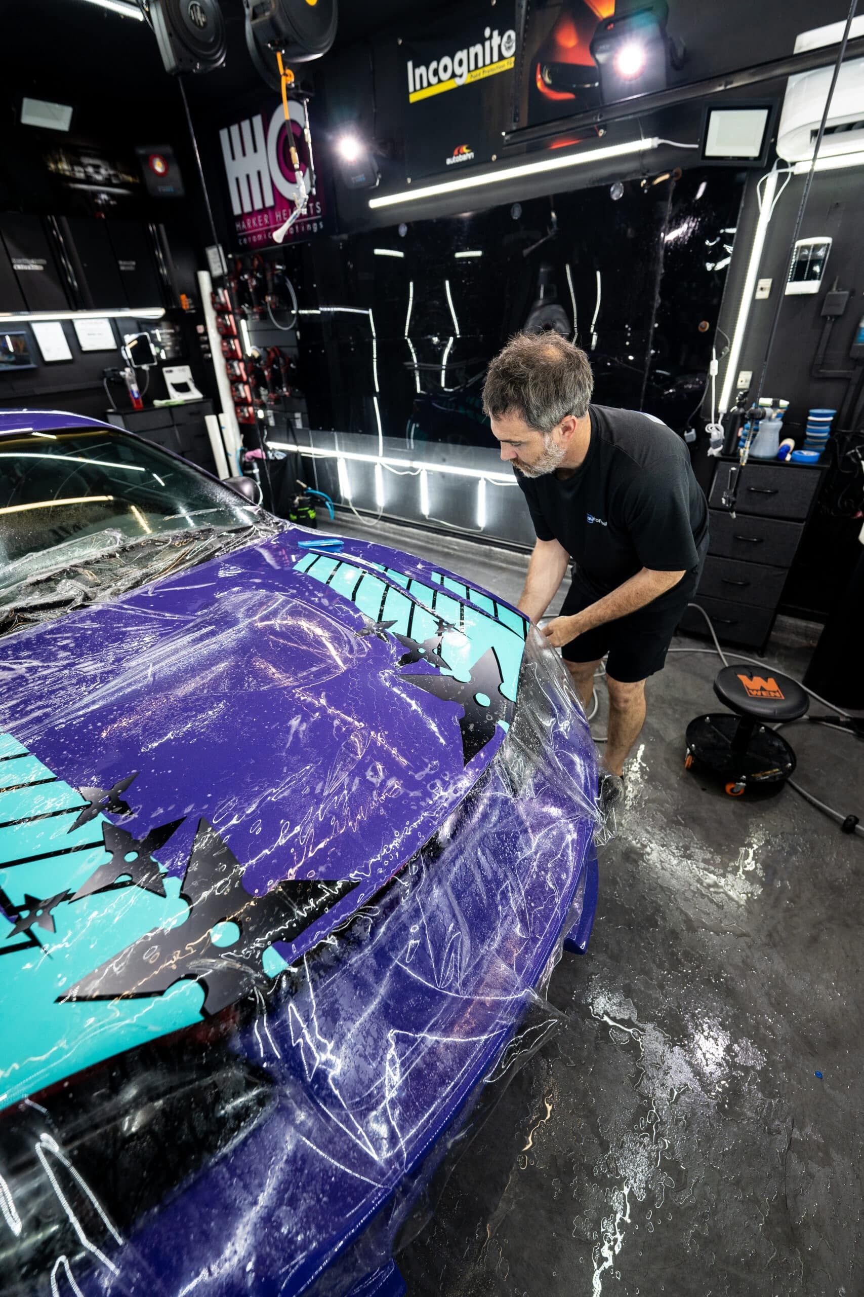 A man is covering a purple car with plastic wrap in a garage.