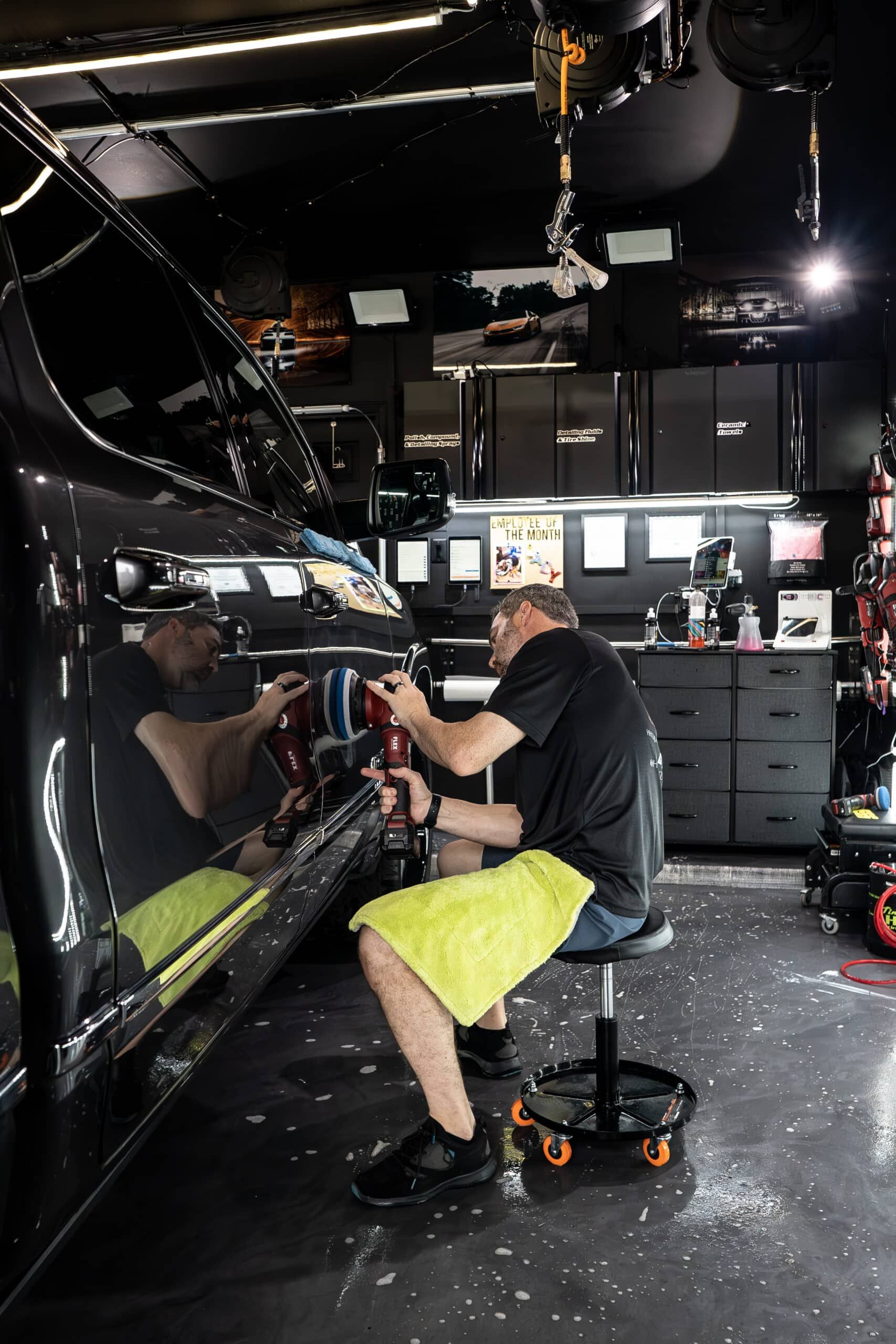 A man is sitting on a stool in a garage polishing a car.
