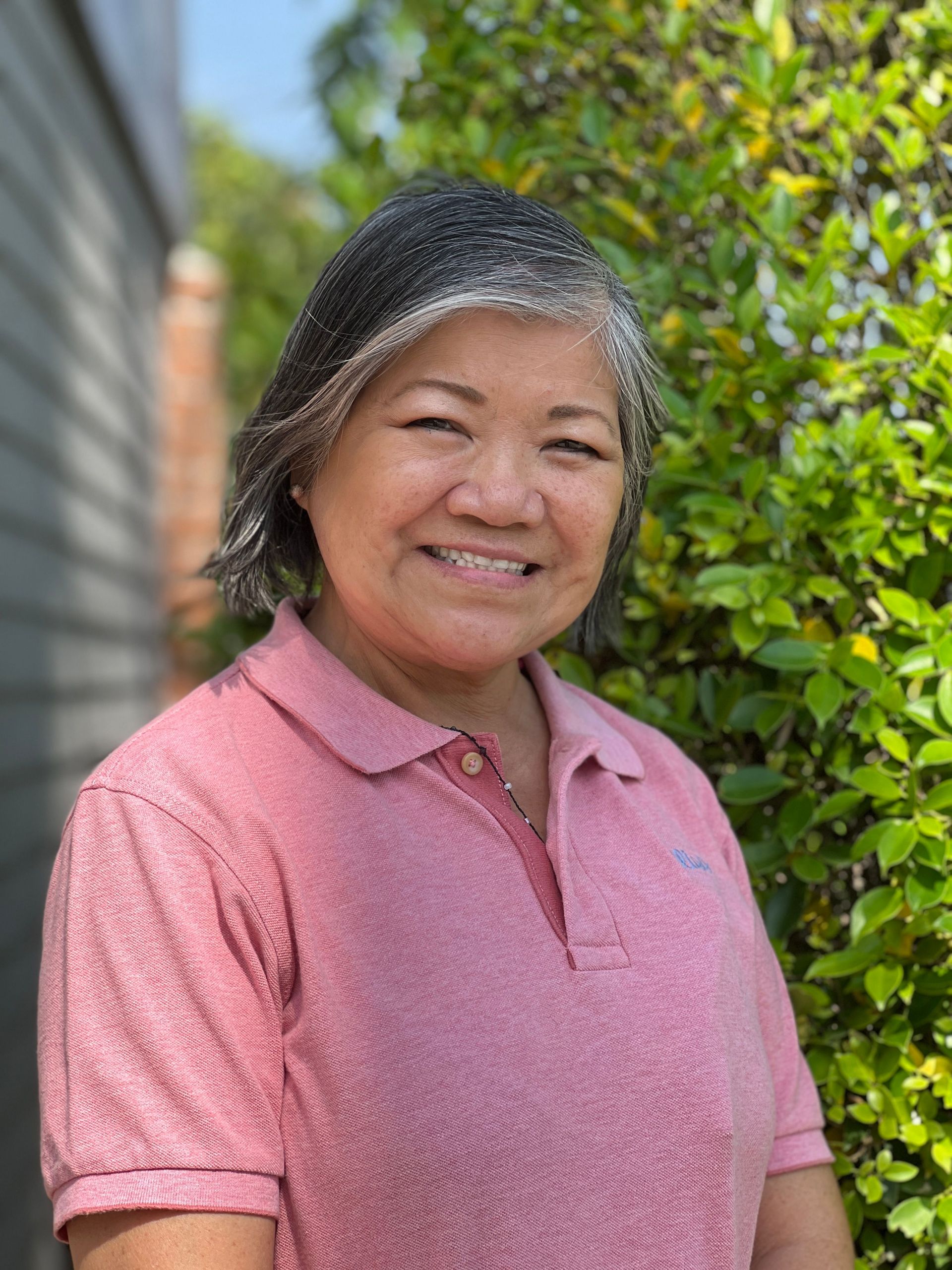 A woman in a pink shirt is smiling in front of a bush.