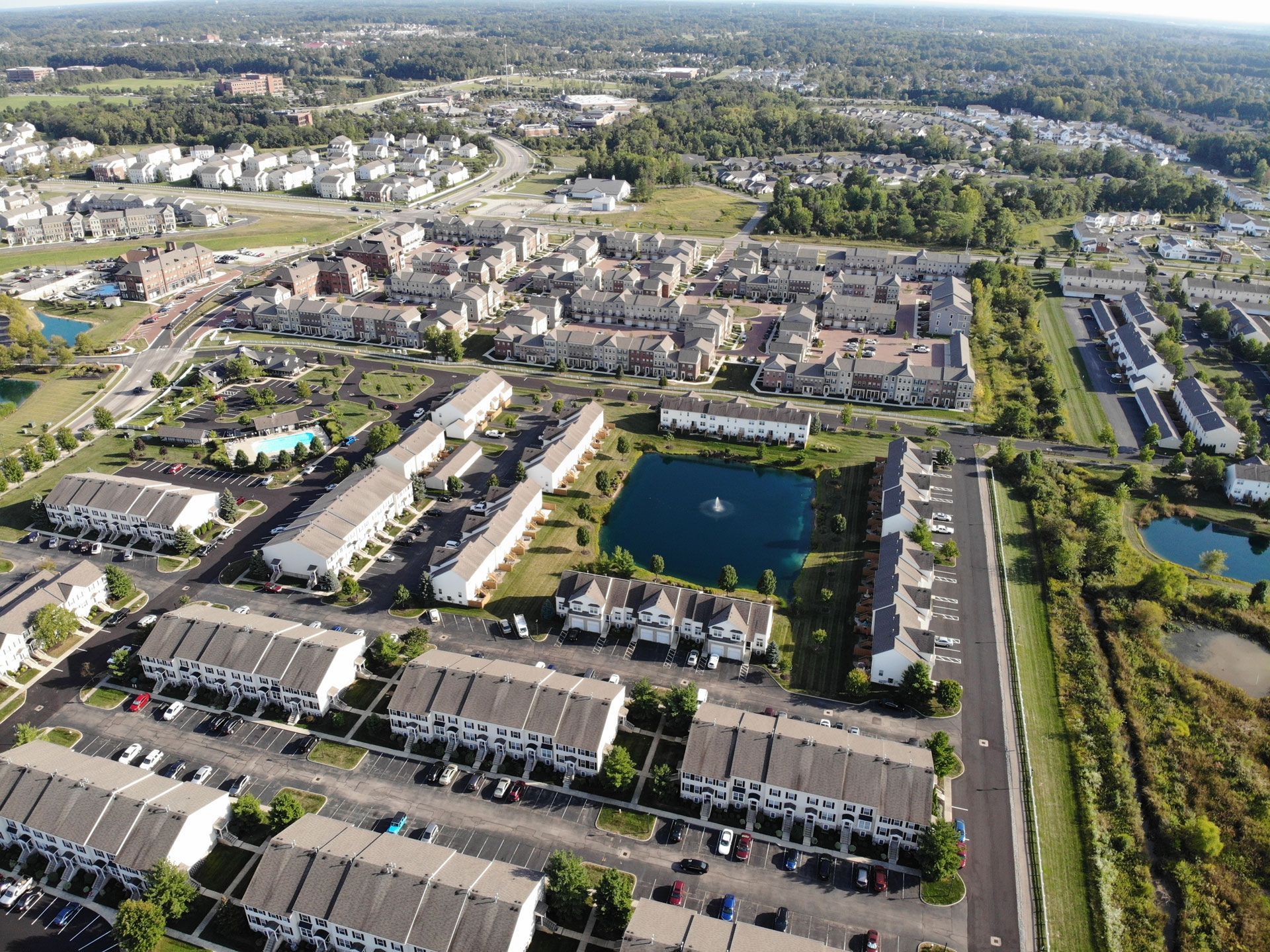 Aerial view of a suburban neighborhood with townhouses, a pond, trees, and parked cars.