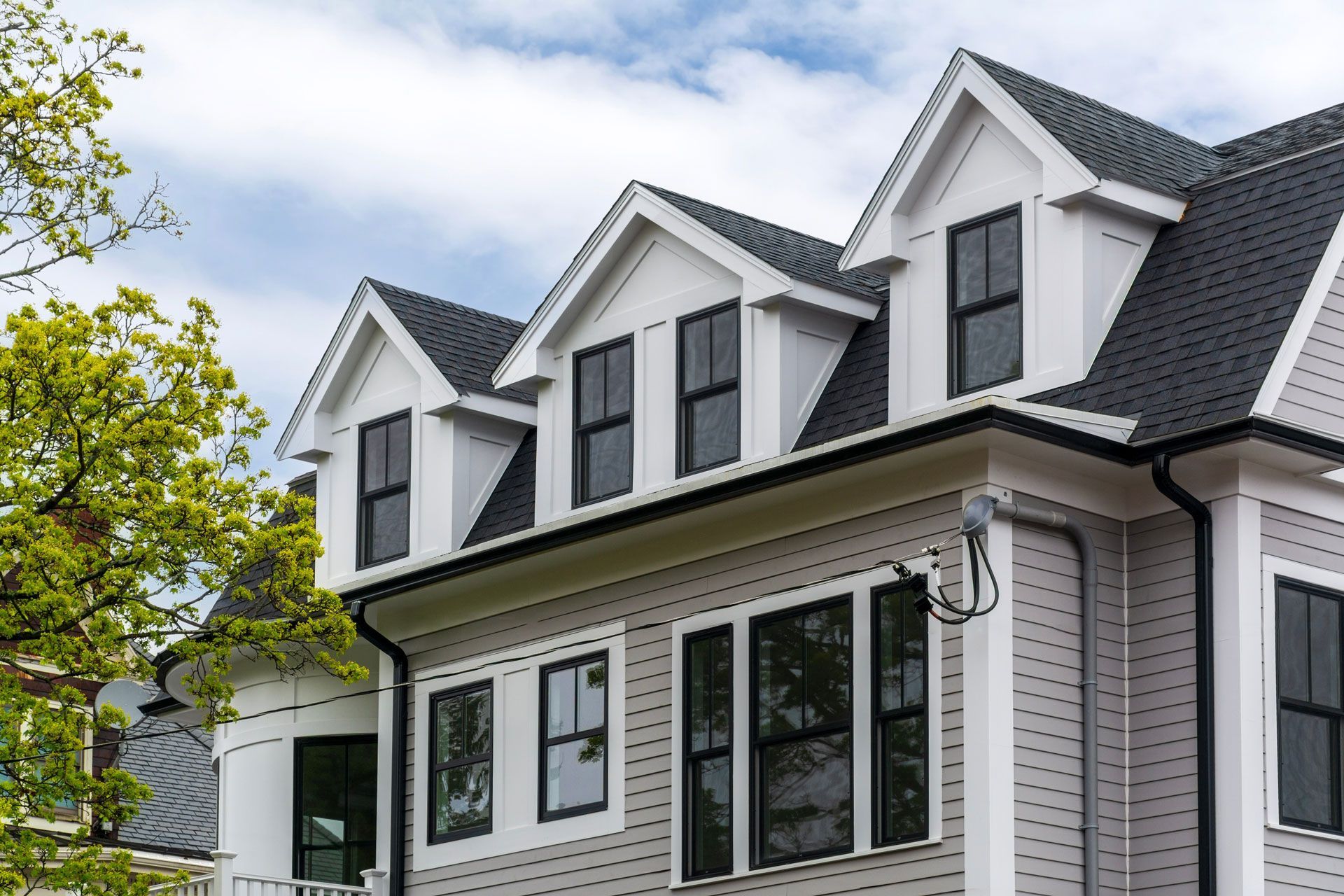 Gray house with three dormers, black windows, and white trim against a cloudy sky.