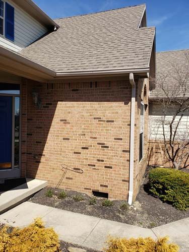 Brick exterior of a house with brown roof, beige gutters, and a small bush.