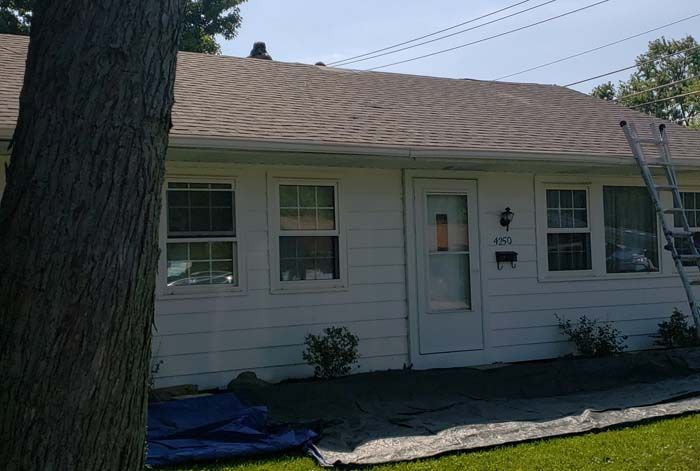 White house with brown roof; ladder leans against it. A tree stands in front, sunny day.