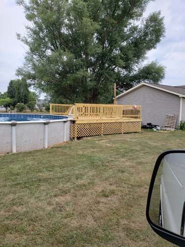 Above ground pool with wooden deck, grass, and a tree in the background.