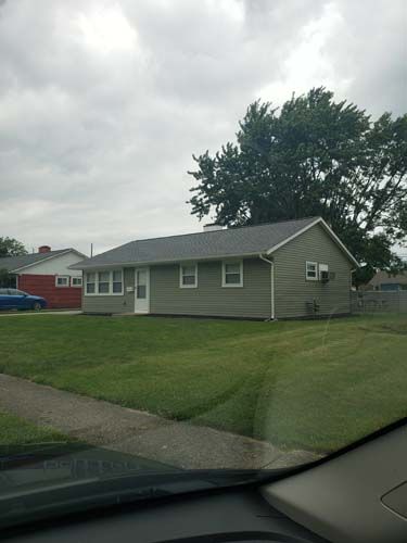 Green-sided ranch house with white trim on a cloudy day; lawn and driveway in front.