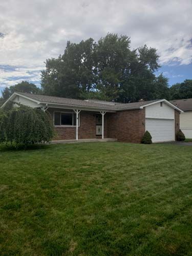 Brick ranch-style house with a garage, surrounded by green grass and trees under a cloudy sky.