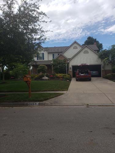 Two-story house with tan siding, a garage, and a red vehicle on the driveway; surrounded by a green lawn and trees.