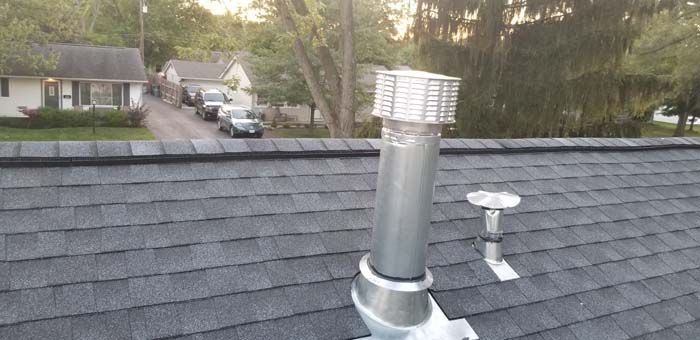 A metal chimney pipe on a gray shingle roof, with houses and trees in the background.