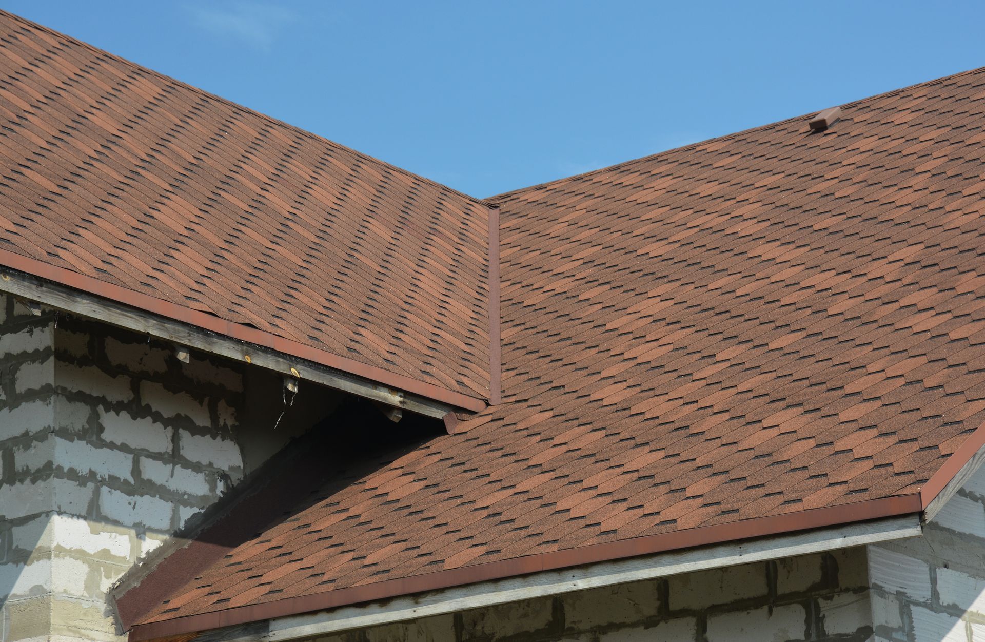 Brown shingled roof with a corner, resting on a light-colored brick building, against a clear blue sky.