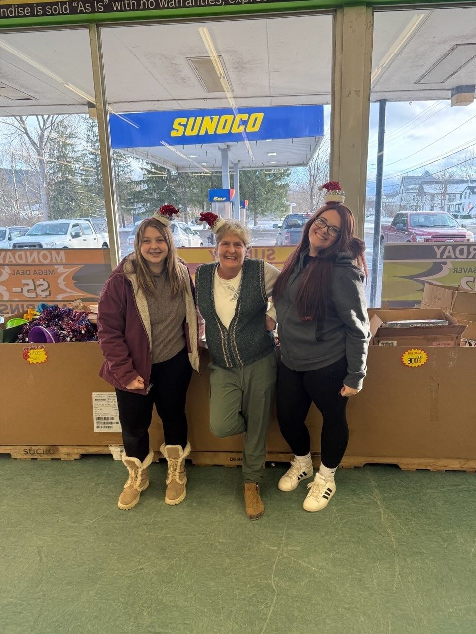 Three people stand smiling in a store with a Sunoco sign outside. They wear casual clothes.