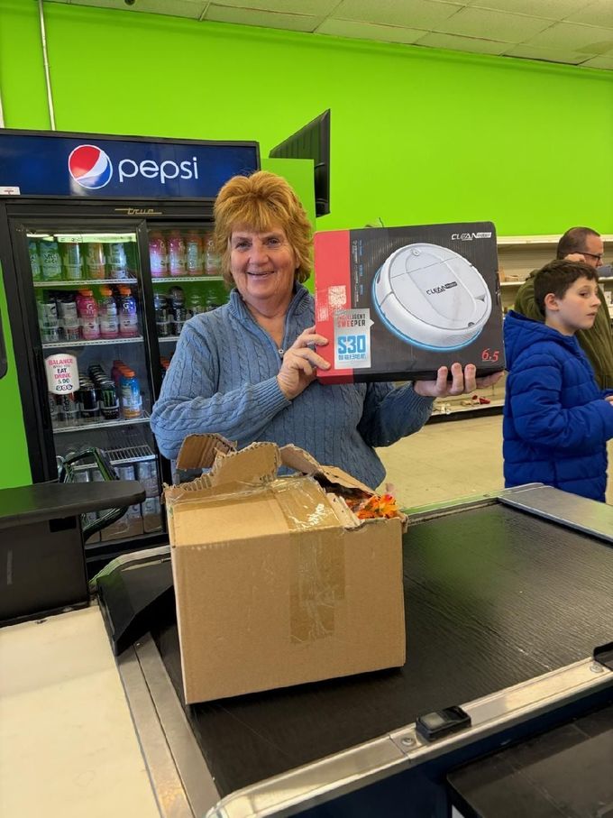 Woman holding a robot vacuum box at a checkout counter, smiles. A child is nearby. Bright green wall.