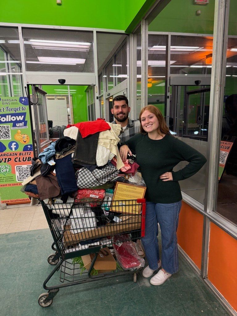 Two people stand next to a shopping cart overflowing with clothing in a brightly lit store.