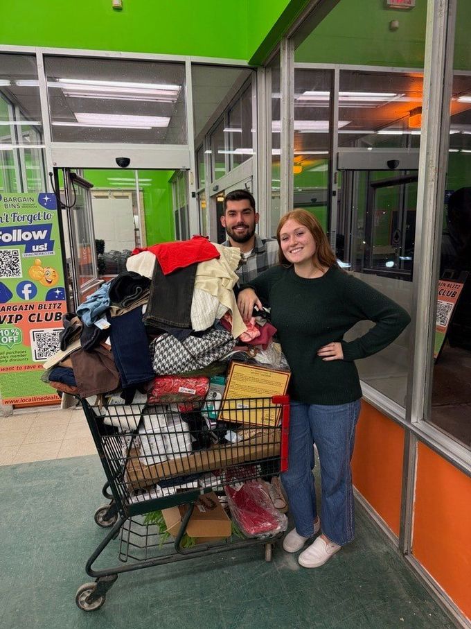 Two people stand next to a shopping cart overflowing with clothing in a brightly lit store.