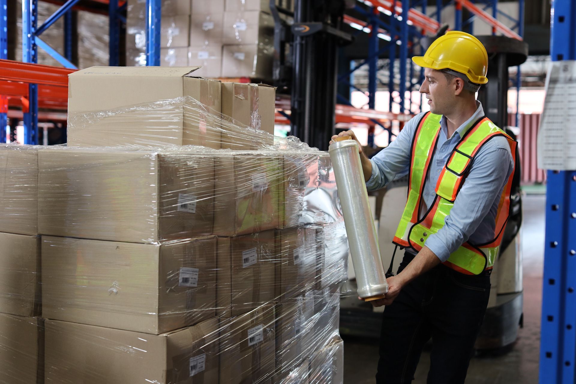A man is wrapping boxes in plastic wrap in a warehouse.