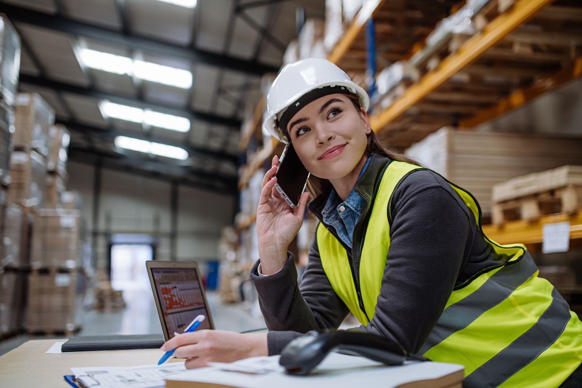 A woman is sitting at a desk in a warehouse talking on a cell phone.