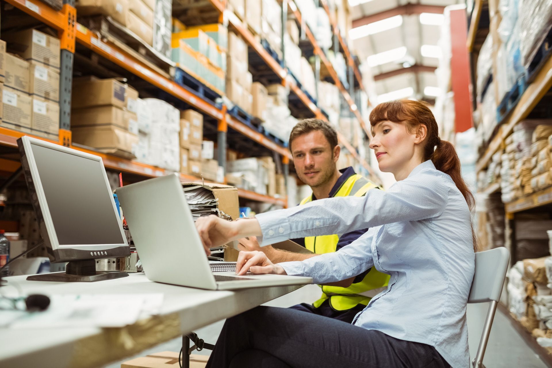 A man and a woman are working on a laptop in a warehouse.