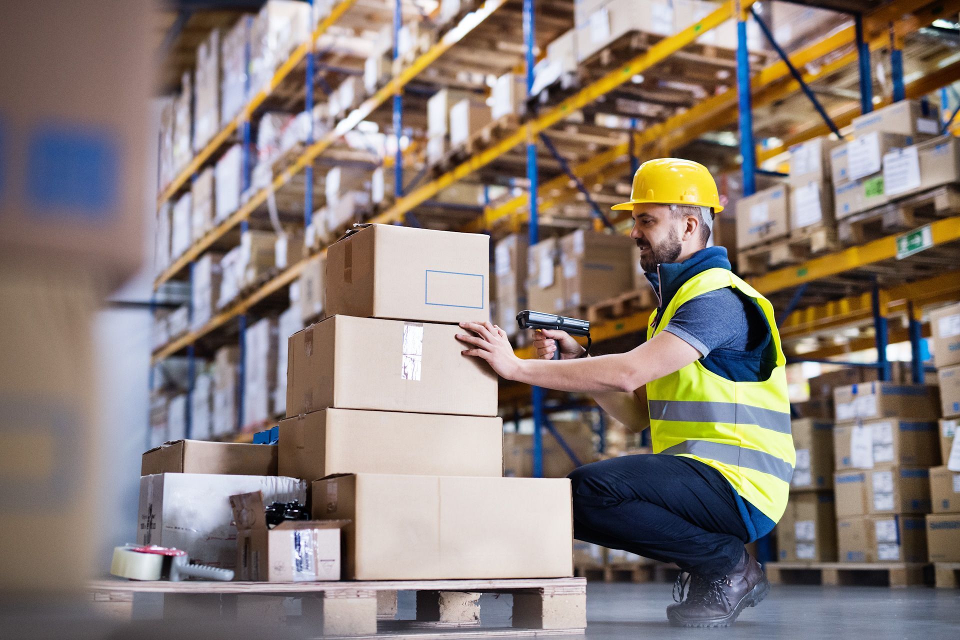A man is kneeling down in a warehouse while using a scanner to scan boxes.