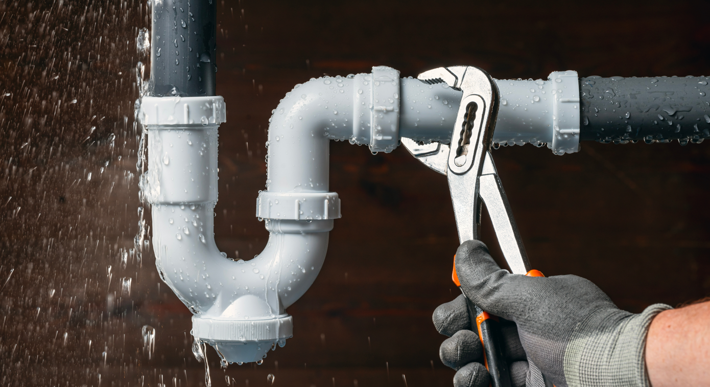 Person with gloved hand uses wrench on leaking sink pipe. Water sprays.