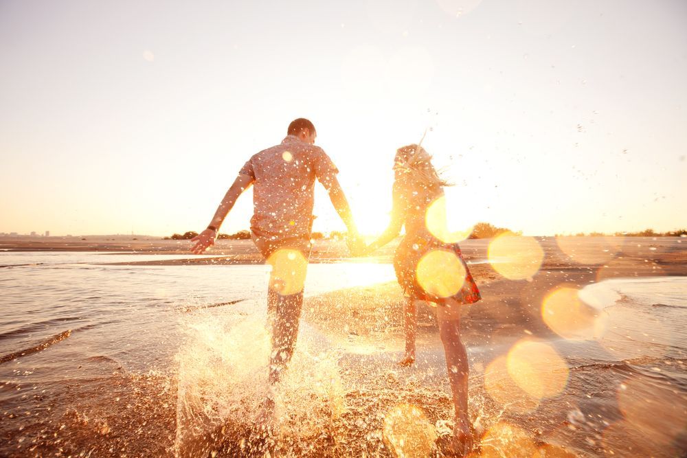 Couple runs, holding hands, through splashing water on a sunny beach at sunset — Bespoke Legal Group In Northern, NSW