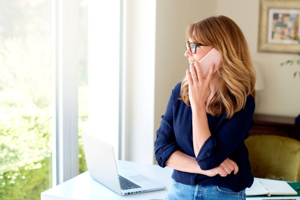 Woman on a phone near a window, holding a laptop. She wears glasses and a dark blue shirt  — Bespoke Legal Group In Southport, QLD