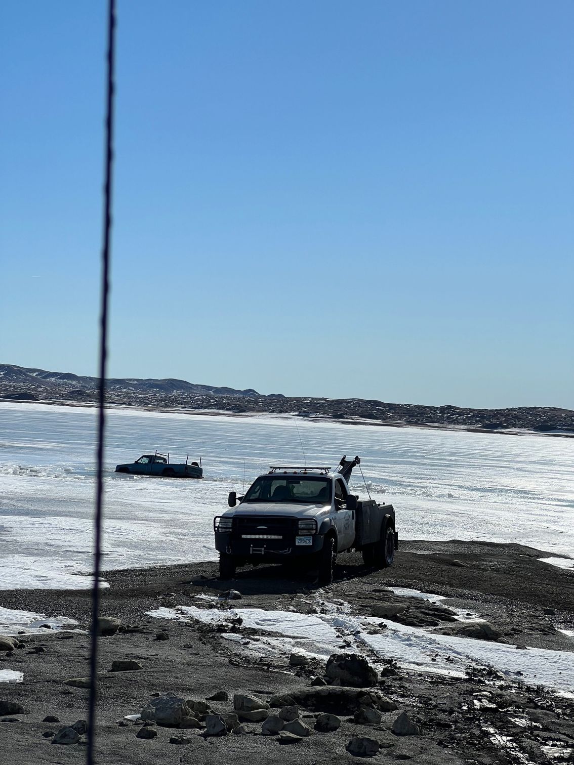 White pickup truck on rocky ground, with a boat in the icy water under a bright blue sky.