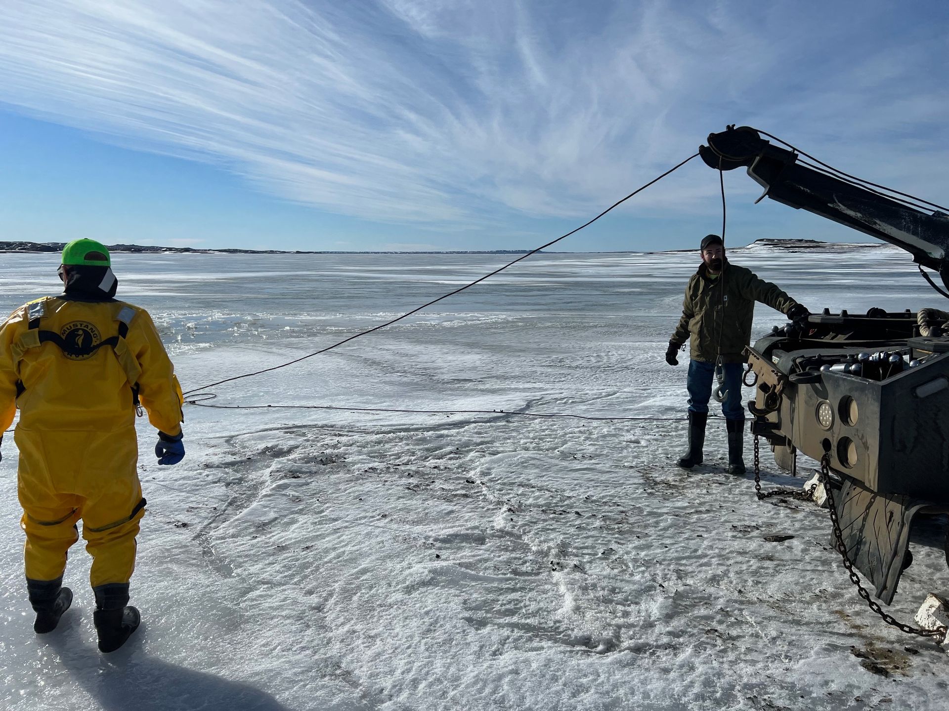 Two people on icy surface, one in yellow suit, operating equipment attached by cable. Sunny day, open water in background.