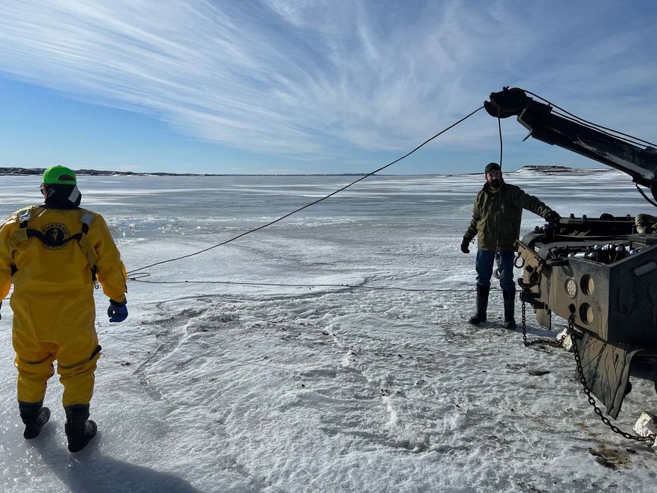 Two people on ice using a winch. One in yellow suit, green helmet; other in a jacket. Clear sky, frozen lake.