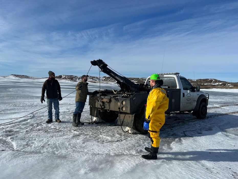 Tow truck on icy ground, pulling something. Three people nearby. Sunny, outdoor setting.