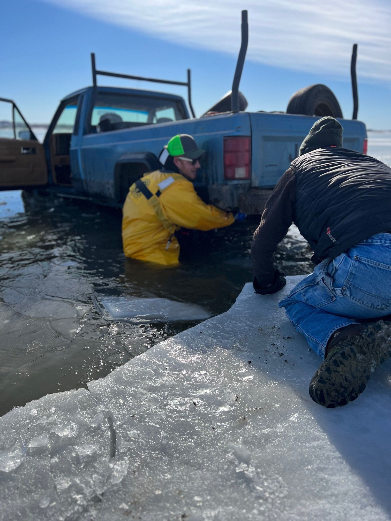 Two people attempt to free a blue truck submerged in icy water; one wears a yellow coat, the other jeans.