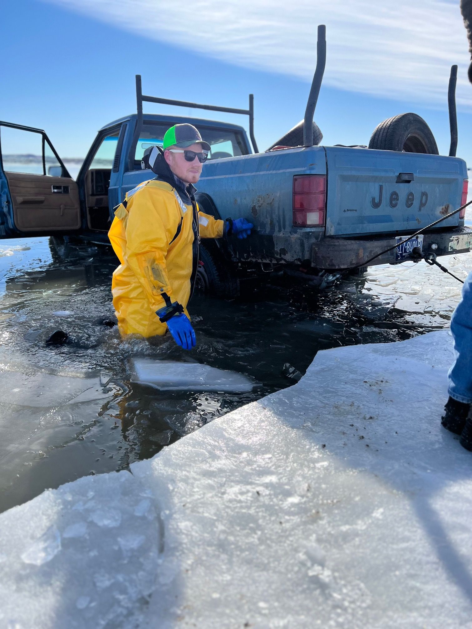 Person in yellow suit near a blue Jeep truck stuck in icy water.