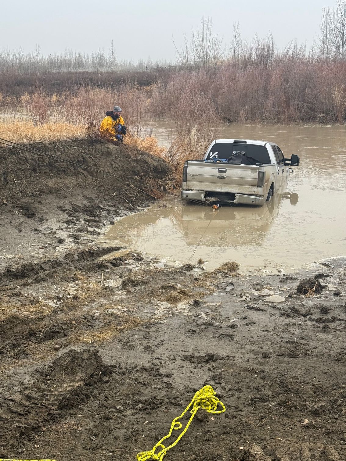 A pickup truck stuck in muddy water. A person in yellow jacket kneels on bank. Yellow tow rope in foreground.