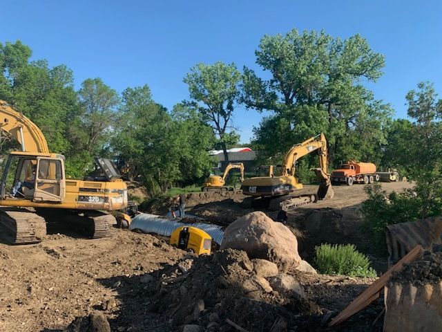 Construction site with multiple yellow excavators digging a trench; trees and blue sky background.