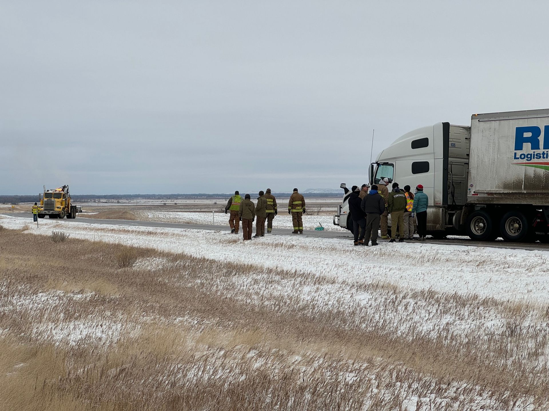 People near a semi-truck in a snowy field. Firefighters and other personnel are present.