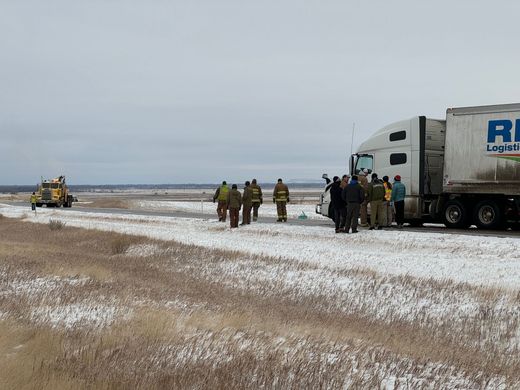 Group of people near semi-truck in snowy field; wrecker in distance.