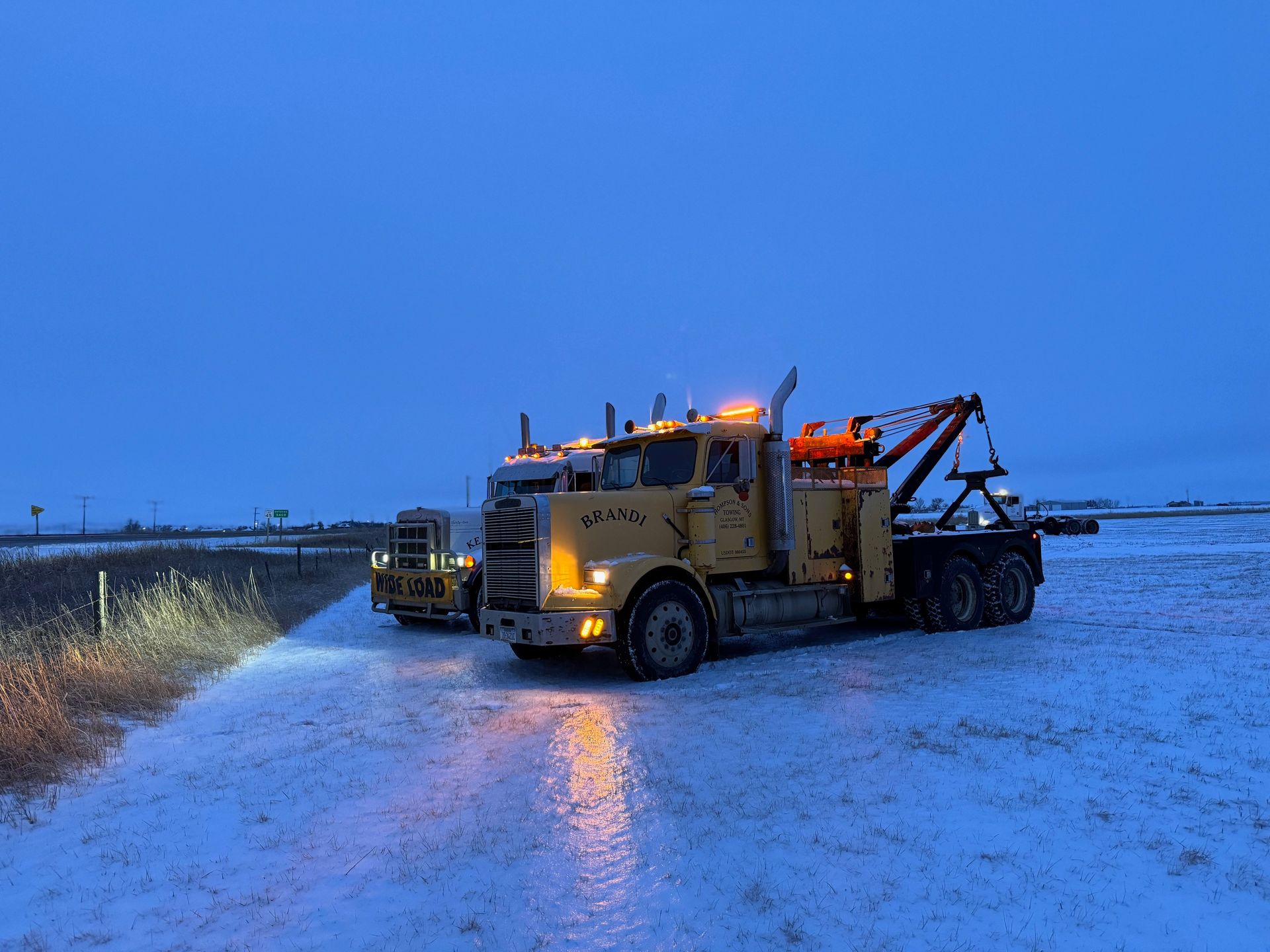 Two yellow tow trucks parked on a snowy road at dusk.