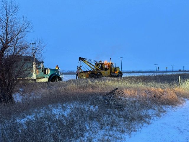 Tow truck pulling a semi-truck out of a snowy ditch alongside a rural road at dusk.